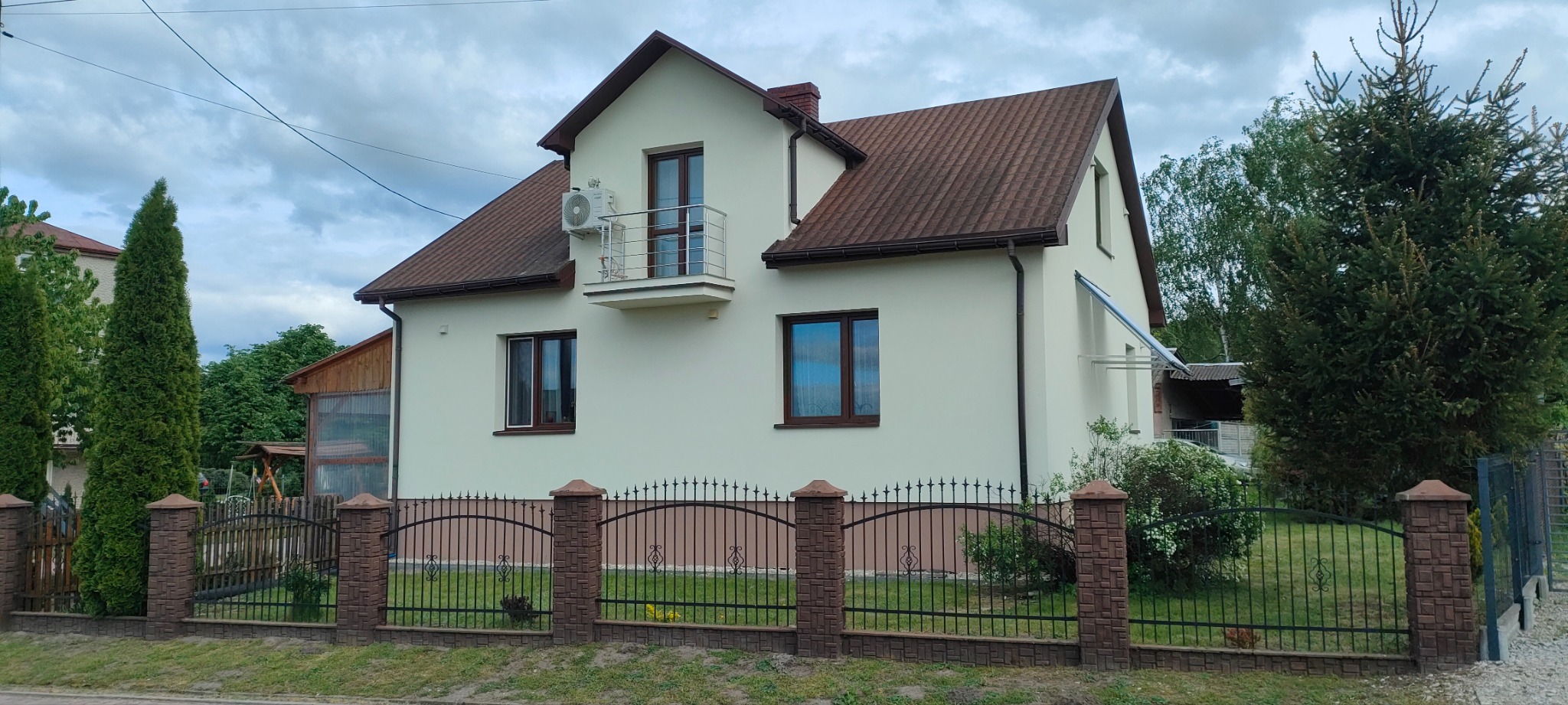 Dom jednorodzinny z elewacją w kolorze jasnego beżu, brązowym dachem i oknami. Widoczny balkon z metalową balustradą oraz klimatyzator na ścianie. Ogród z metalowym ogrodzeniem.
