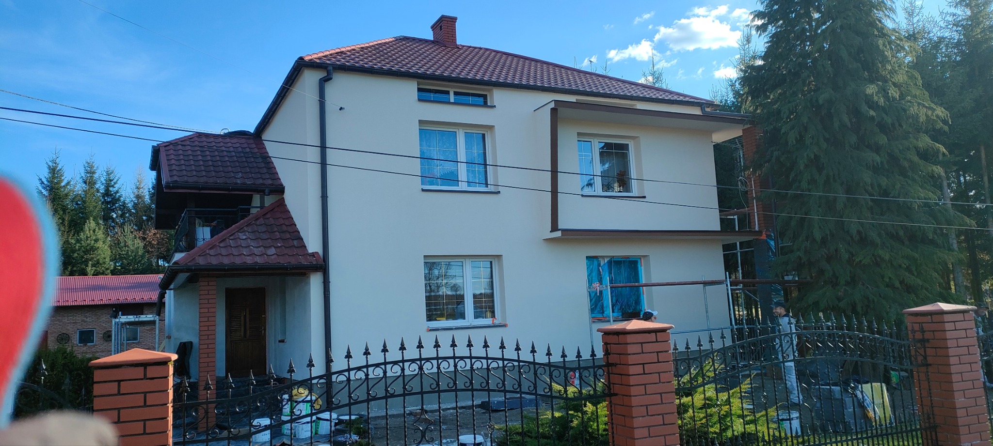 Dom po remoncie elewacji w Górze, widoczny przez ozdobne ogrodzenie. Świeżo pomalowana fasada w jasnym kolorze, z ciemnobrązowymi detalami. Częściowo widoczne rusztowania.