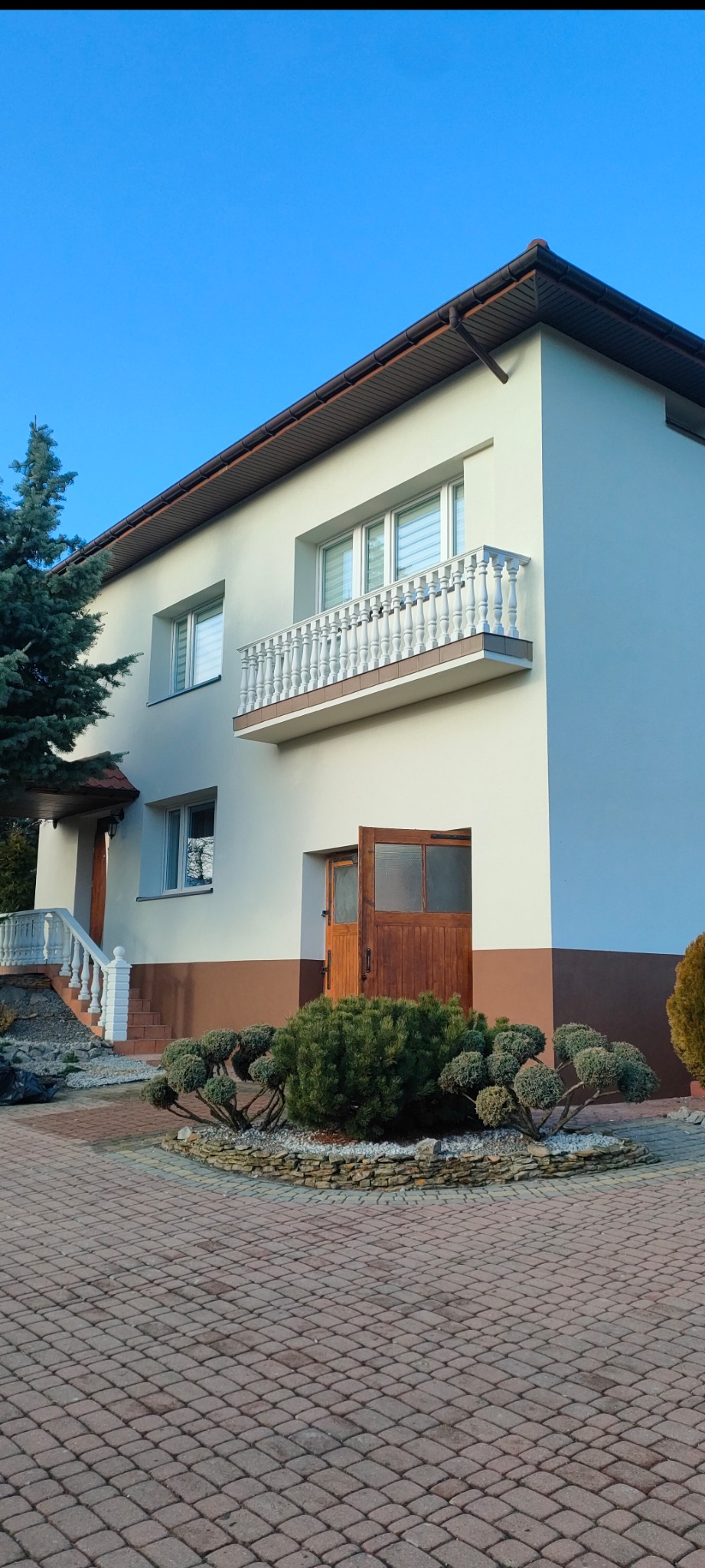 Elewacja dwukondygnacyjnego domu z balkonem i drewnianymi drzwiami wejściowymi, wykończona w jasnych kolorach z brązowym pasem u dołu.