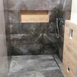 Kabina prysznicowa z szarymi płytkami imitującymi marmur, drewniana wnęka w ścianie i odpływ liniowy. Widoczny chromowany zestaw natryskowy.