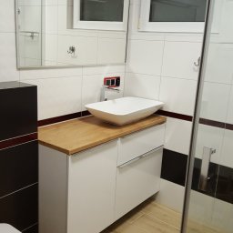 Usługi glazurnicze - Nowoczesna łazienka z białą umywalką nablatową na szafce z drewnianym blatem, jasne płytki na ścianach i podłodze imitujące drewno, kabina prysznicowa z szybą.