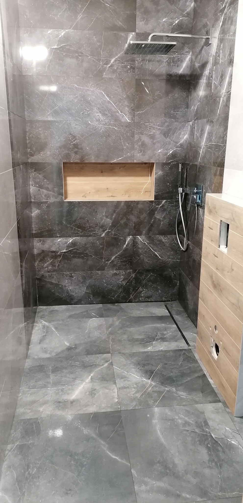 Kabina prysznicowa z szarymi płytkami imitującymi marmur, drewniana wnęka w ścianie i odpływ liniowy. Widoczny chromowany zestaw natryskowy.