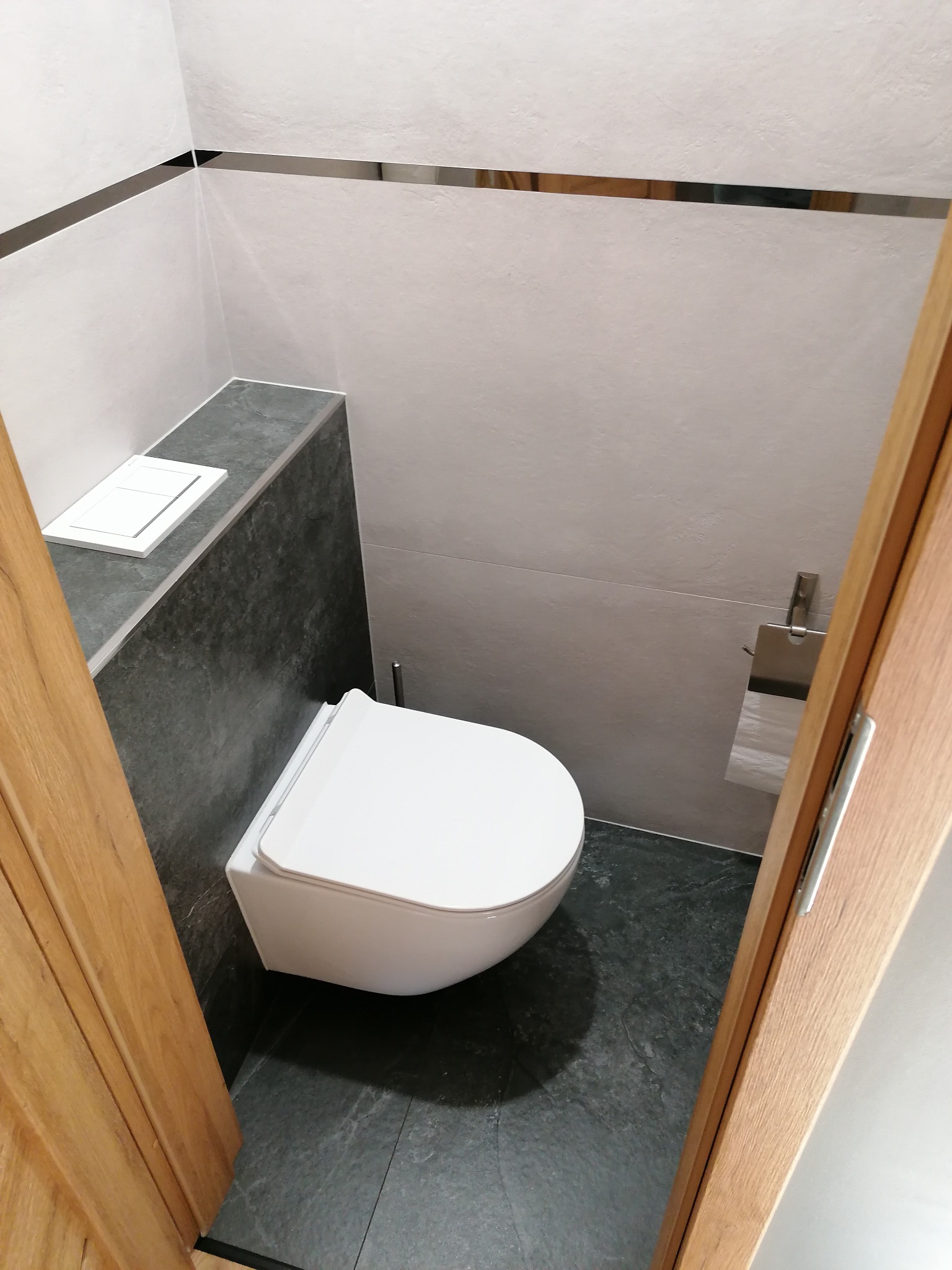 Nowoczesna toaleta podwieszana w łazience wykończonej szarymi płytkami i elementami dekoracyjnymi ze stali nierdzewnej. Widok zza drewnianej ramy drzwi.