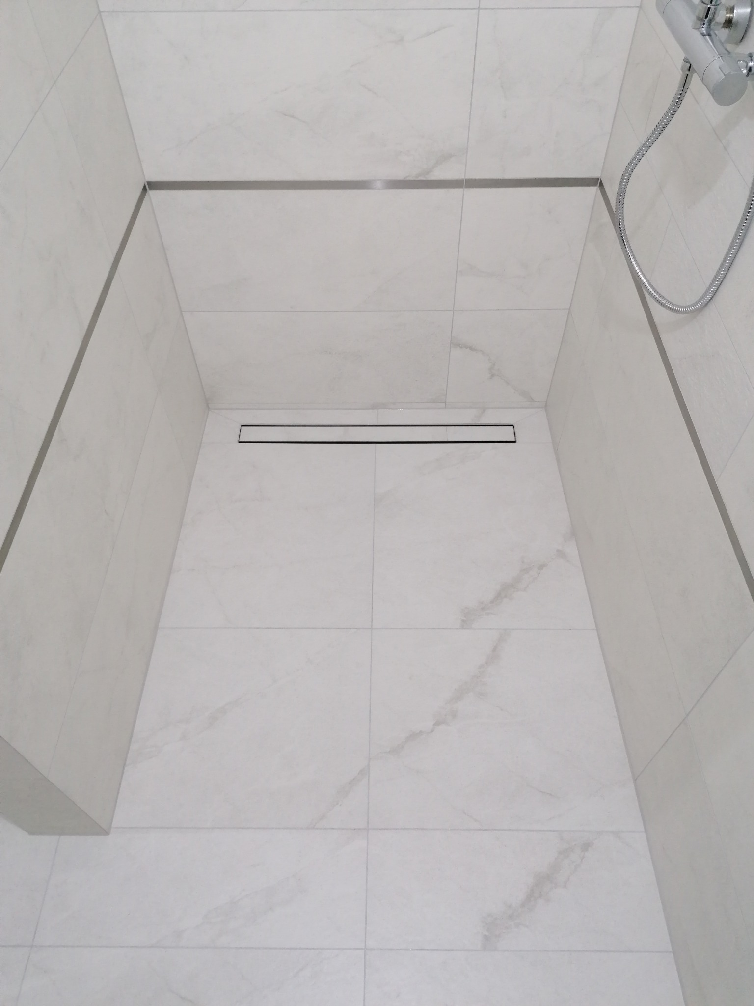 Nowoczesny brodzik wyłożony białymi płytkami imitującymi marmur, z liniowym odpływem i chromowaną armaturą prysznicową. Minimalistyczny design.