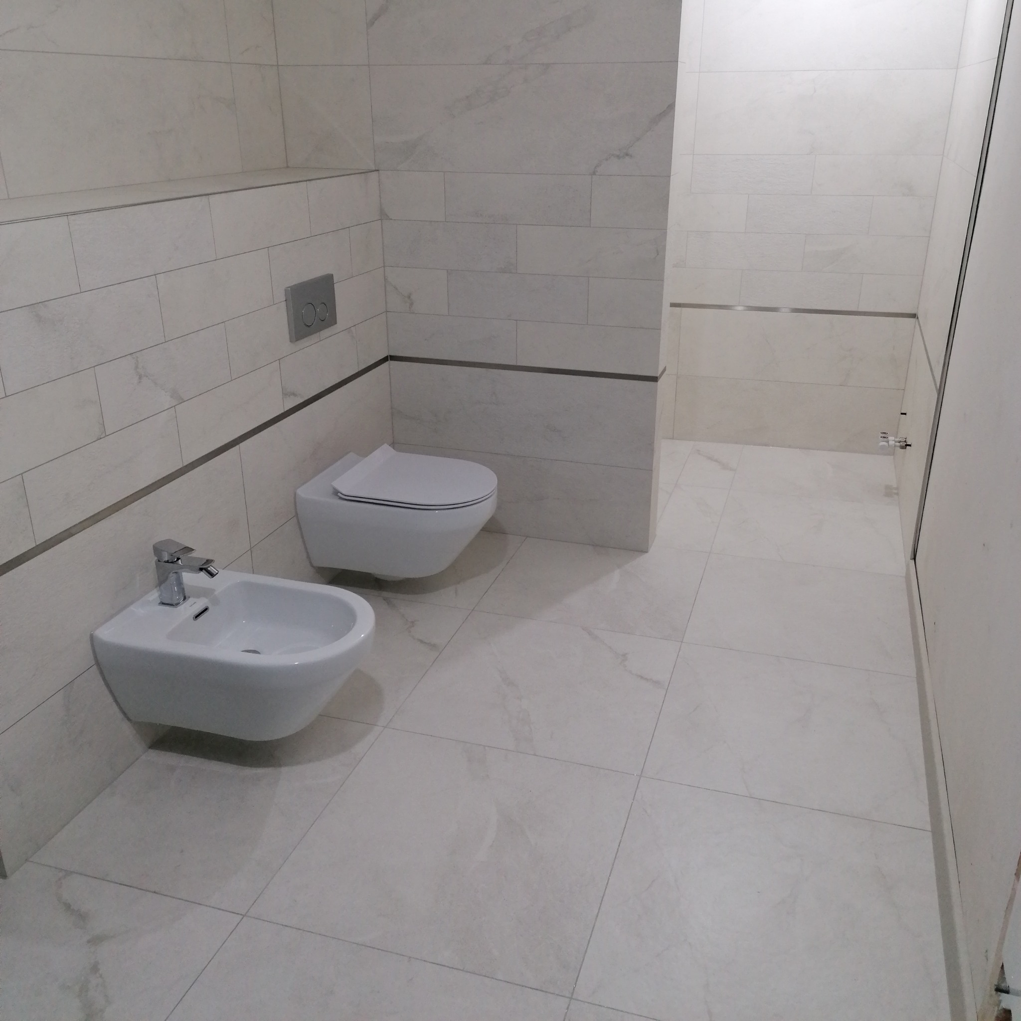 Jasna łazienka z beżowymi płytkami, wiszącą toaletą i bidetem. Widoczne srebrne listwy dekoracyjne i przycisk spłuczki. Minimalistyczny wystrój, nowoczesne sanitariaty.