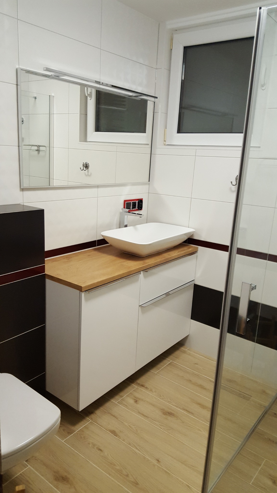 Nowoczesna łazienka z białą umywalką nablatową na szafce z drewnianym blatem, jasne płytki na ścianach i podłodze imitujące drewno, kabina prysznicowa z szybą.