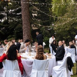 SHEF-POVAR CATERING SPÓŁKA Z OGRANICZONĄ ODPOWIEDZIALNOŚCIĄ - Uroczystość na świeżym powietrzu: goście siedzą przy okrągłych stołach z białymi obrusami, dekorowanych świecami, pod girlandami światełek, w otoczeniu zieleni.