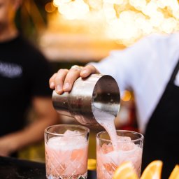 SHEF-POVAR CATERING SPÓŁKA Z OGRANICZONĄ ODPOWIEDZIALNOŚCIĄ - Barman nalewa różowy koktajl z shakera do szklanek z lodem na błyszczącym barze, dekoracja z pomarańczy, w tle rozmyte światła.
