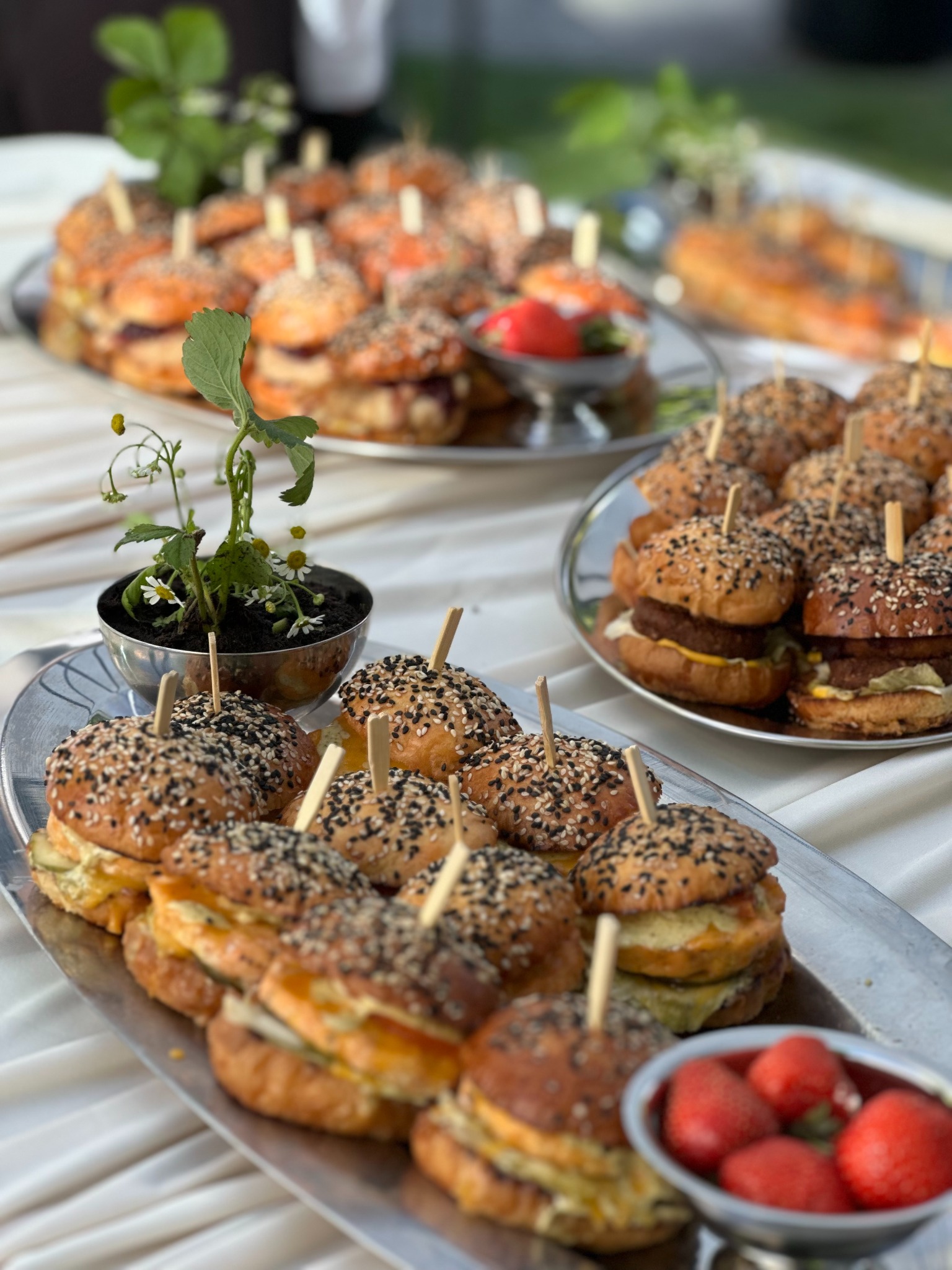 Elegancko podane mini burgery z sezamem na srebrnych tacach, dekorowane roślinami w srebrnych miseczkach i truskawkami, catering na przyjęciu.