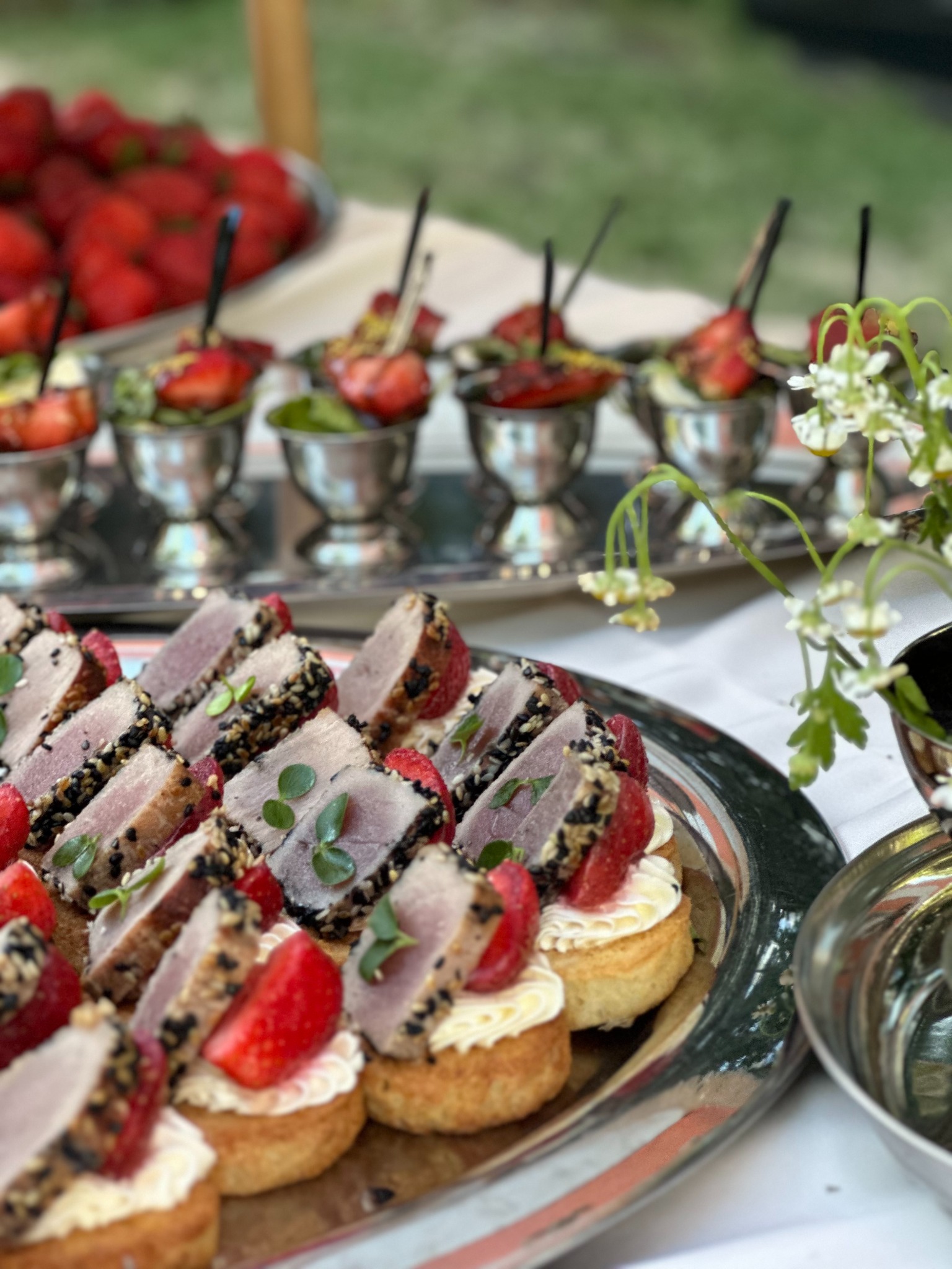 Elegancko podane przekąski cateringowe: mini kanapeczki z tuńczykiem w sezamie i truskawką na srebrnej tacy, w tle kieliszki z deserami truskawkowymi i kwiaty.