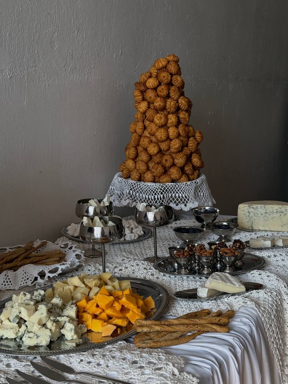 Elegancki stół cateringowy: piramida z ciasta, sery, orzechy, grissini, srebrna zastawa, biały obrus z koronką na tle szarej ściany. Wykwintne przyjęcie.