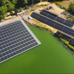 SOLLMAN ADRIAN SACHAJ - Widok z lotu ptaka na pływającą instalację fotowoltaiczną na stawie i panele na lądzie, otoczone zielenią, w słoneczny dzień. Nietypowe rozwiązanie.