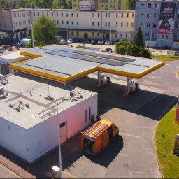 SOLLMAN ADRIAN SACHAJ - Stacja paliw z panelami słonecznymi na dachu w słoneczny dzień. Pomarańczowy samochód serwisowy z drabiną obok budynku stacji. Widok z lotu ptaka.