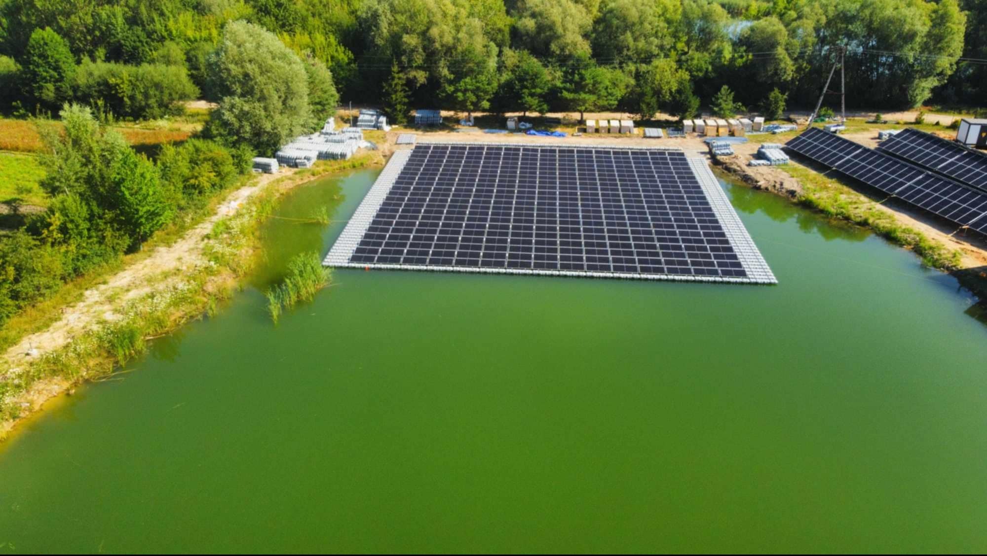 Widok z lotu ptaka na pływającą farmę fotowoltaiczną na jeziorze, z panelami słonecznymi ułożonymi w kwadratową strukturę. Dodatkowe panele na brzegu.