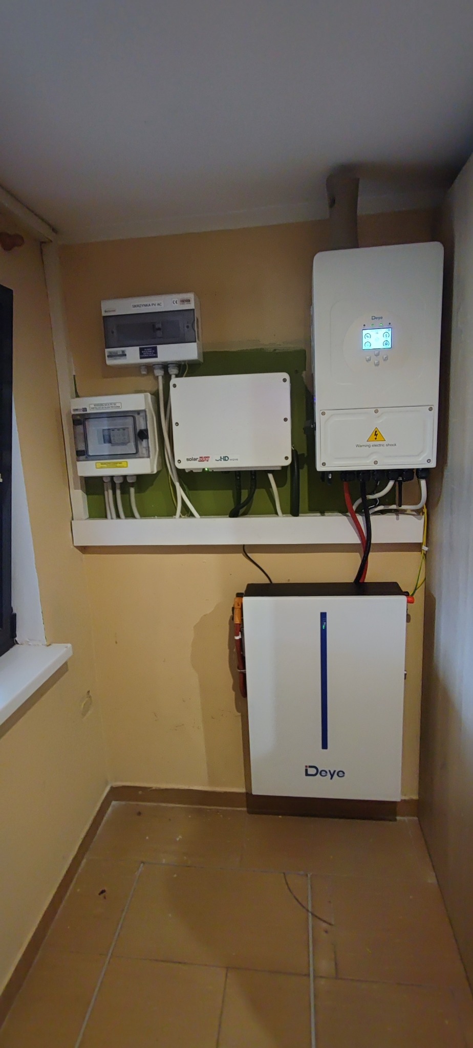 Kompletna instalacja fotowoltaiczna w pomieszczeniu, obejmująca inwerter Deye, magazyn energii, skrzynki AC/DC i optymalizator SolarEdge, zamontowane na ścianie.