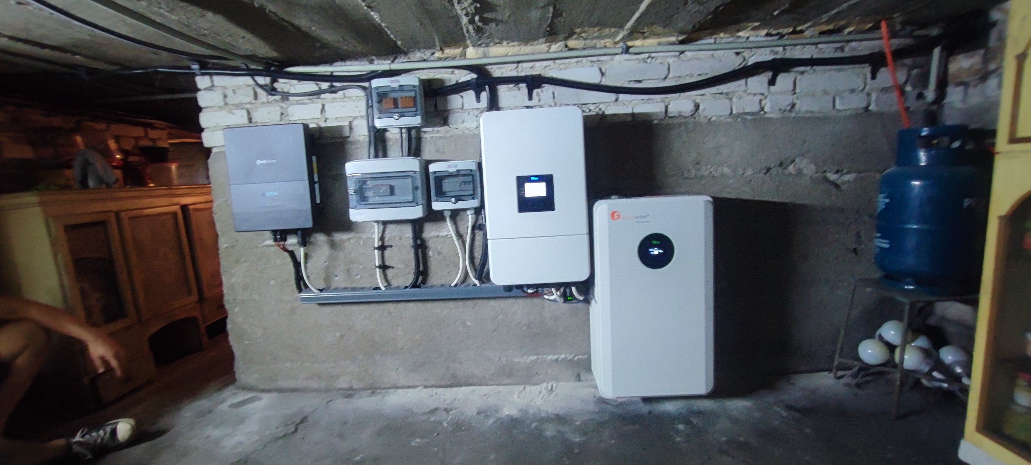 Instalacja paneli fotowoltaicznych w piwnicy: widoczne inwertery, rozdzielnice i akumulator energii. Elementy zamontowane na ścianie z cegły i betonu.