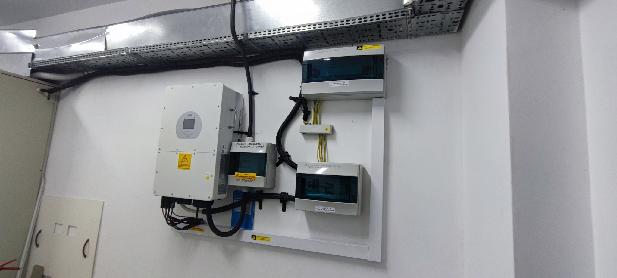 Biały falownik i skrzynki elektryczne zamontowane na ścianie, z widocznymi przewodami i oznaczeniami ostrzegawczymi. Instalacja wewnątrz budynku z metalową korytką kablową.