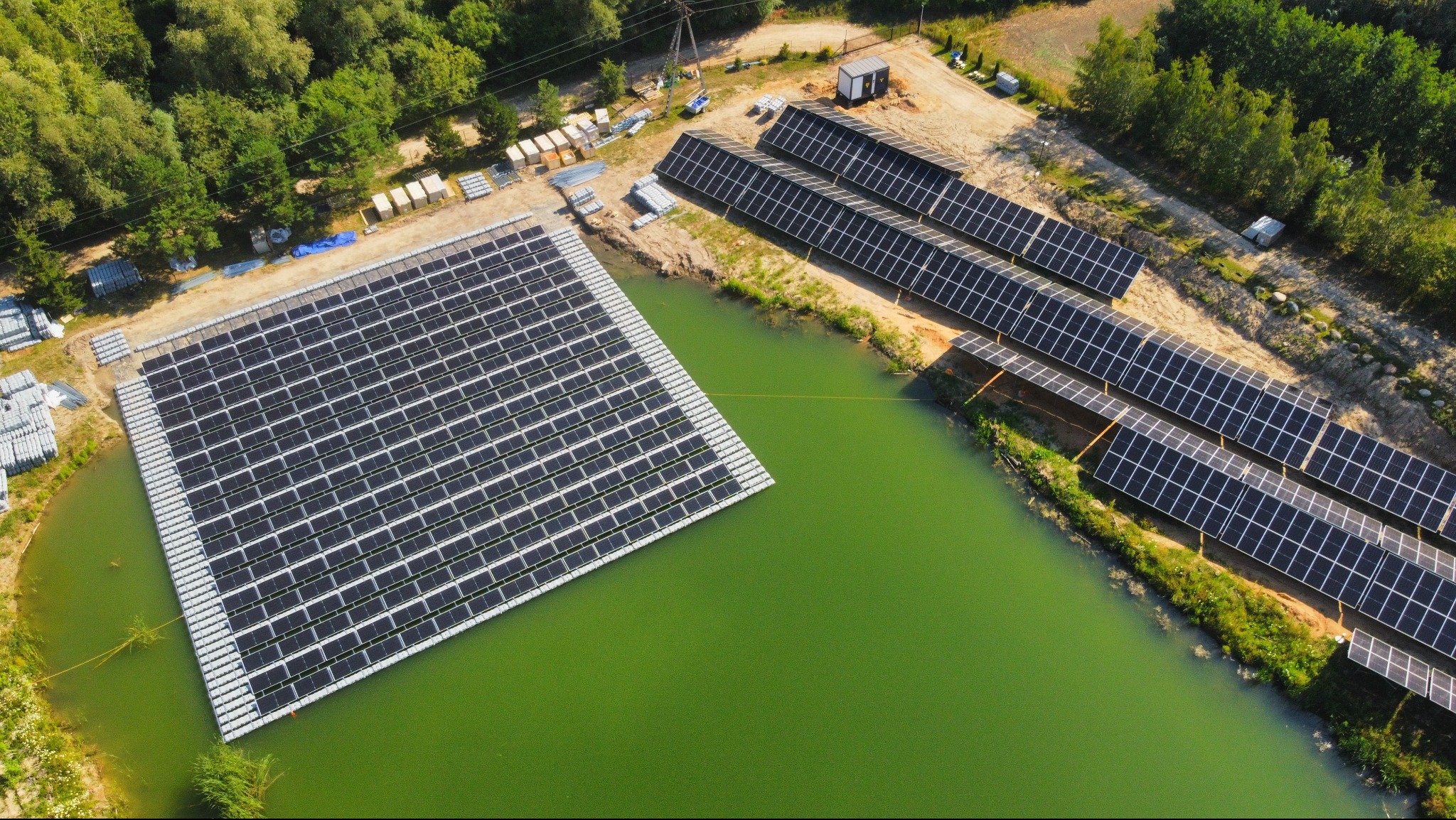 Widok z lotu ptaka na pływającą instalację fotowoltaiczną na stawie i panele na lądzie, otoczone zielenią, w słoneczny dzień. Nietypowe rozwiązanie.
