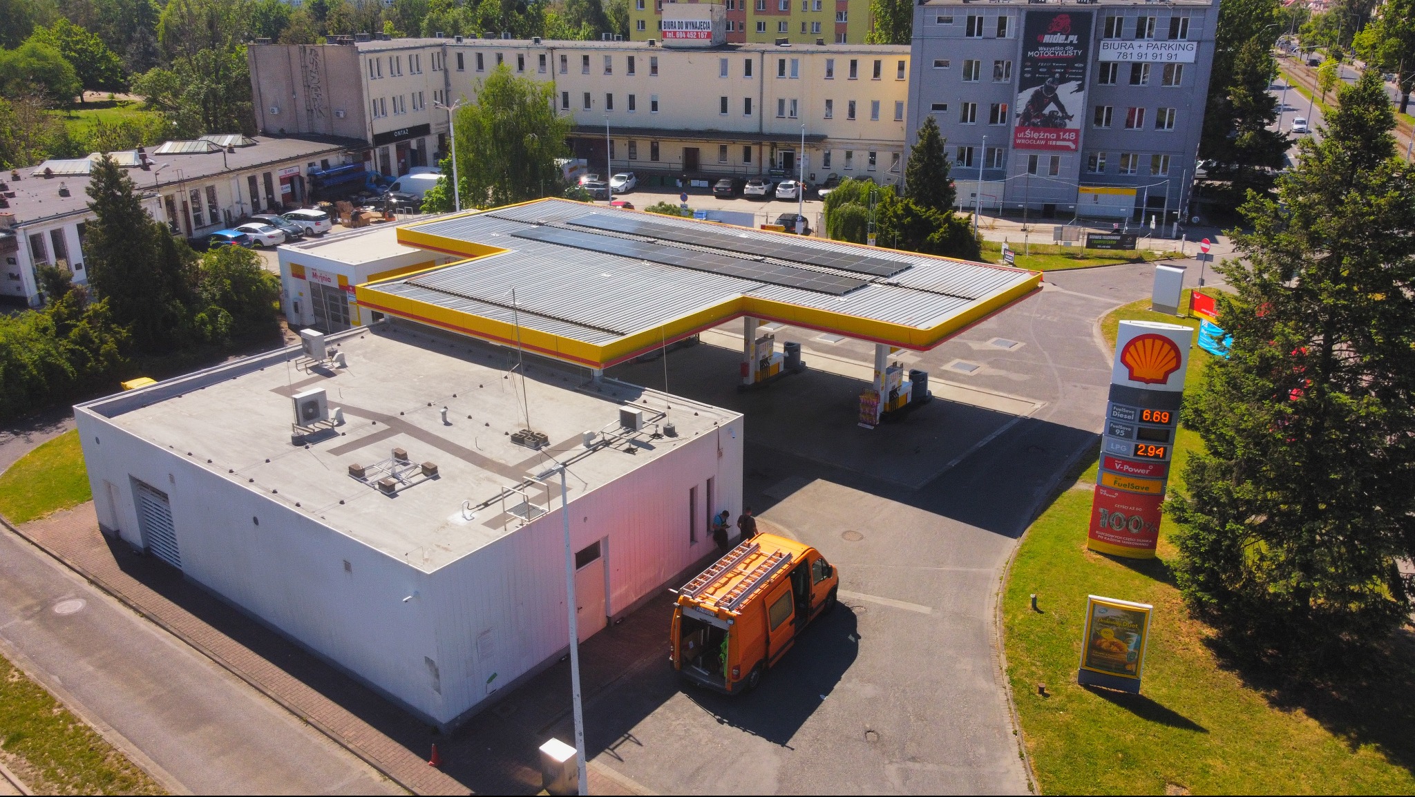 Stacja paliw z panelami słonecznymi na dachu w słoneczny dzień. Pomarańczowy samochód serwisowy z drabiną obok budynku stacji. Widok z lotu ptaka.