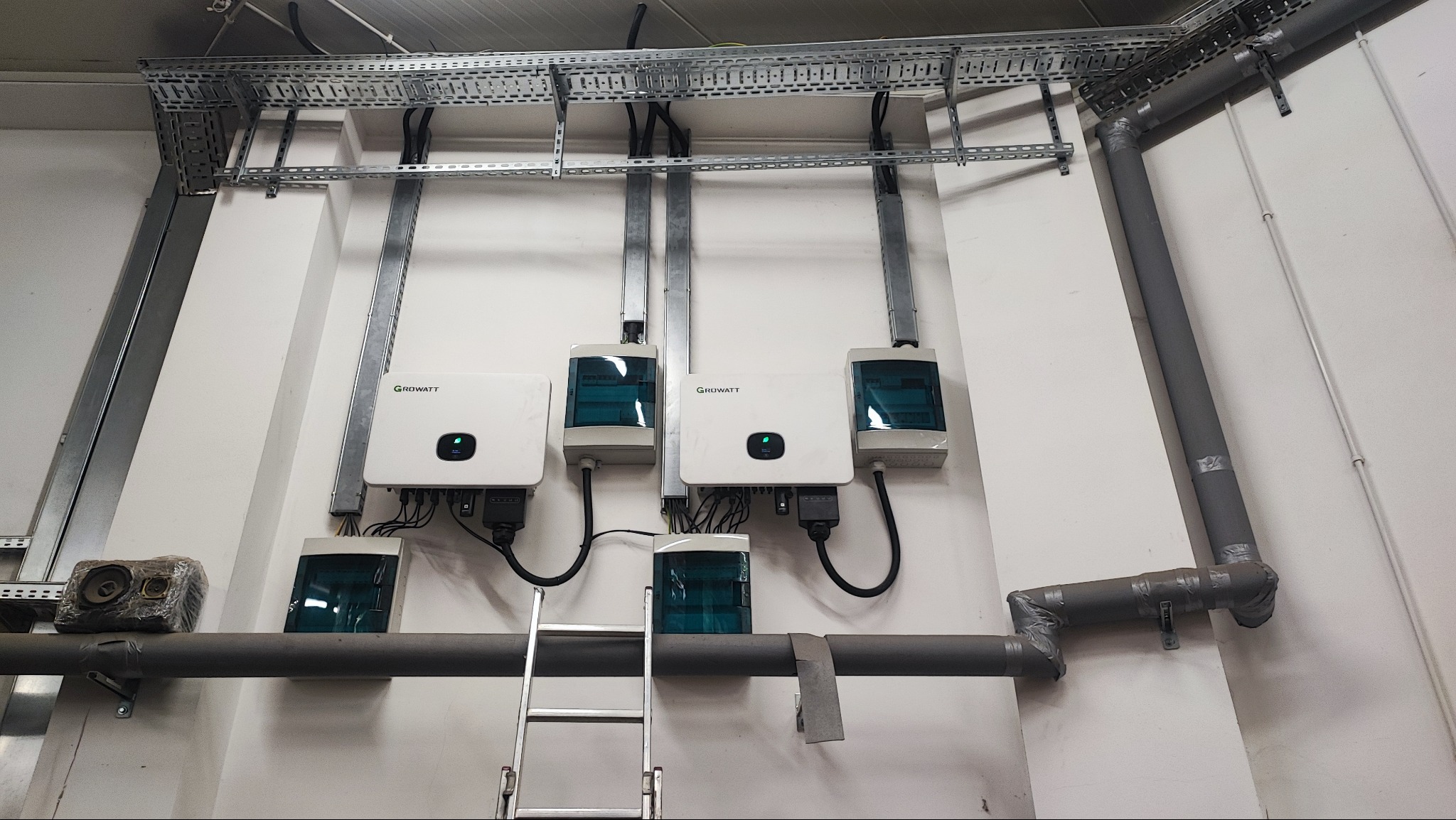 Instalacja trzech białych inwerterów Growatt na ścianie z szarą rurą i drabinką. Metalowe korytka kablowe pod sufitem. Widok z dołu na instalację.