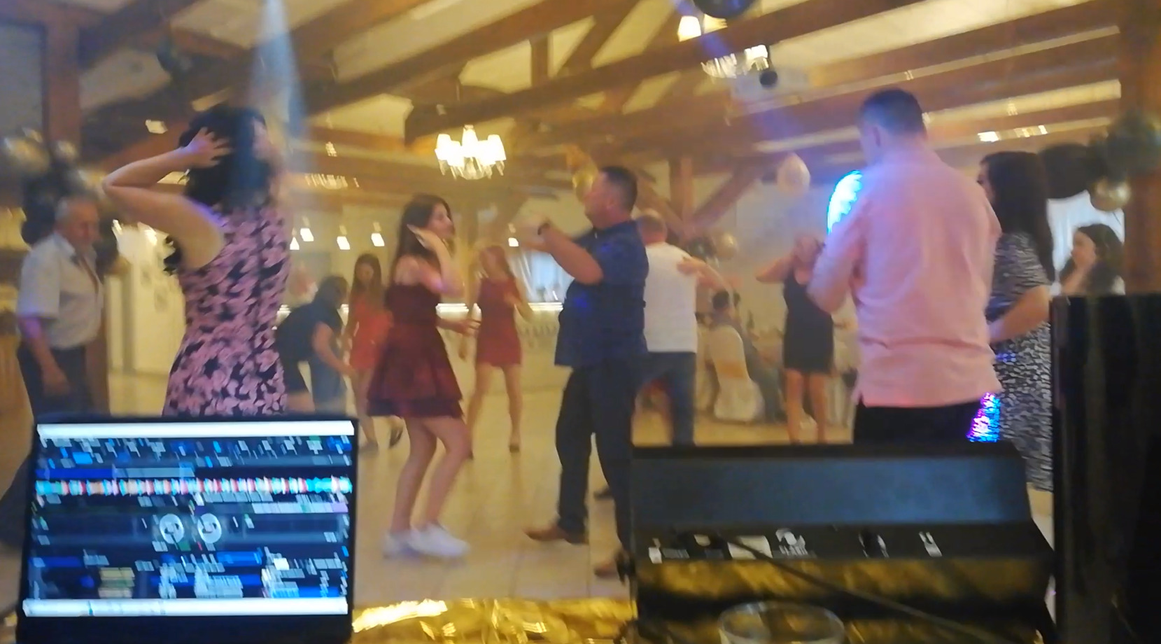 Parkiet taneczny z bawiącymi się gośćmi weselnymi, oświetlony reflektorami. W tle widoczny sprzęt DJ-ski, w tym laptop z oprogramowaniem.