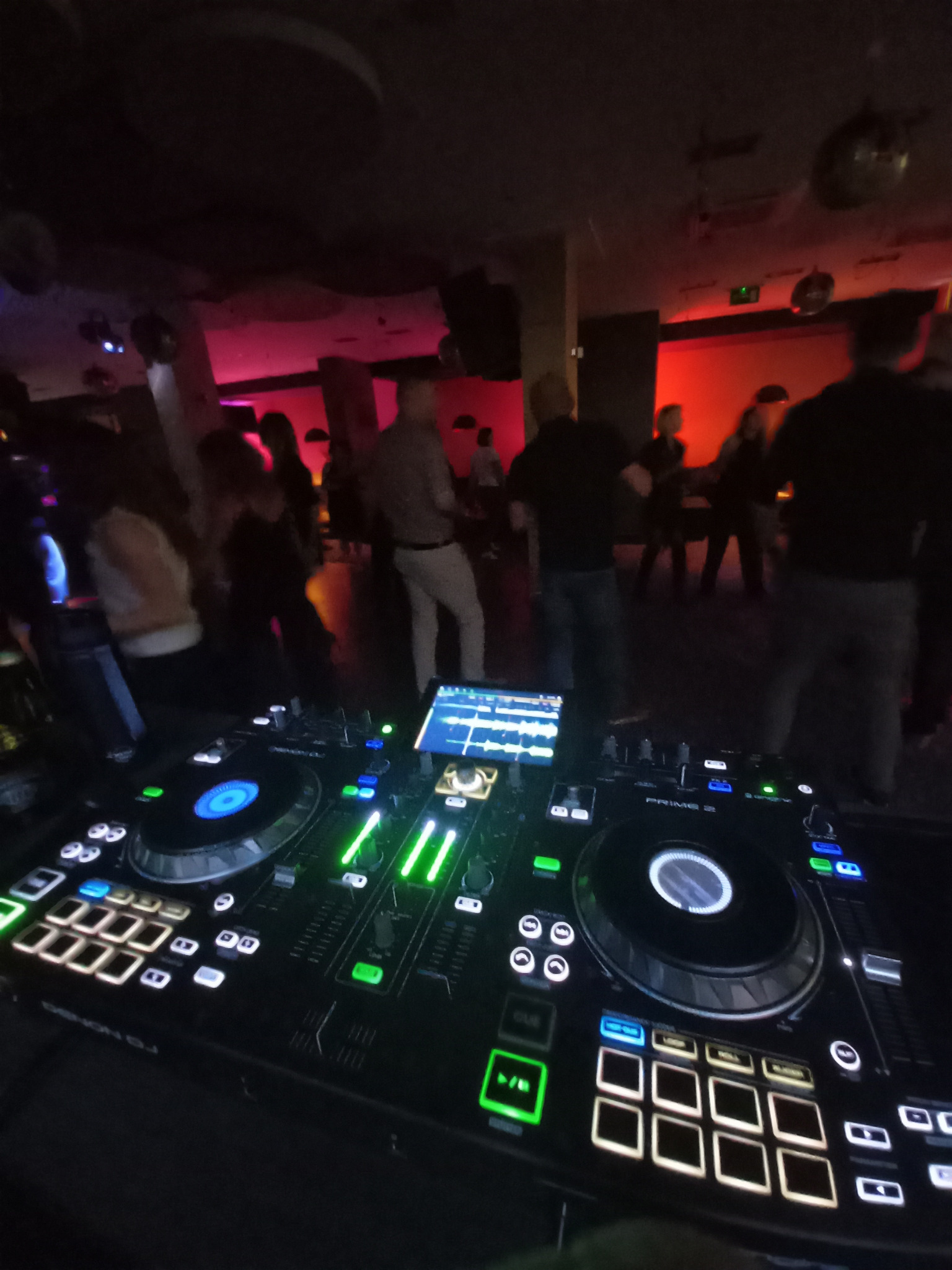 DJ miksuje muzykę na imprezie, w tle tańczący ludzie w czerwonym oświetleniu. Widoczny sprzęt DJ-ski z podświetlanymi przyciskami i ekranem.