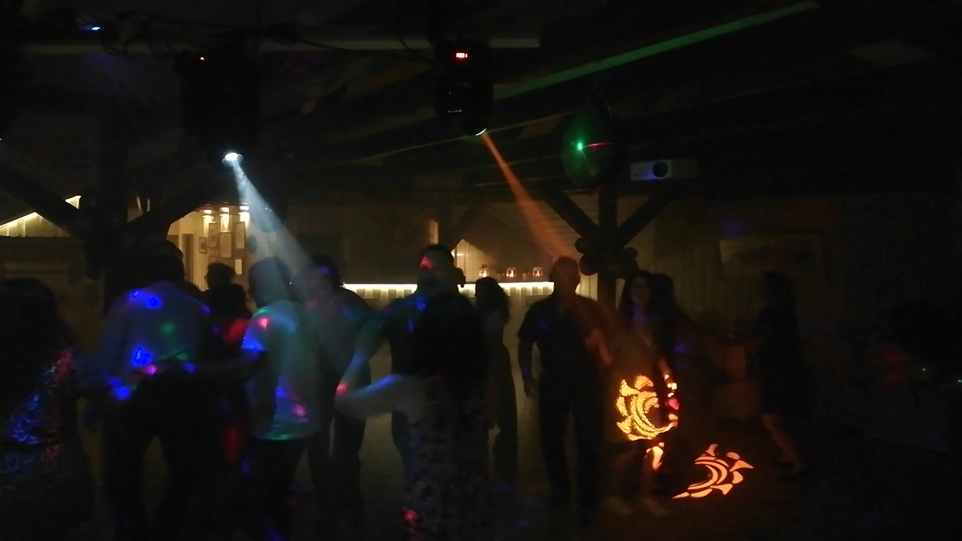 Parkiet taneczny wypełniony bawiącymi się ludźmi, oświetlony dynamicznymi światłami dyskotekowymi i projektorem, tworzącymi wzory na podłodze.