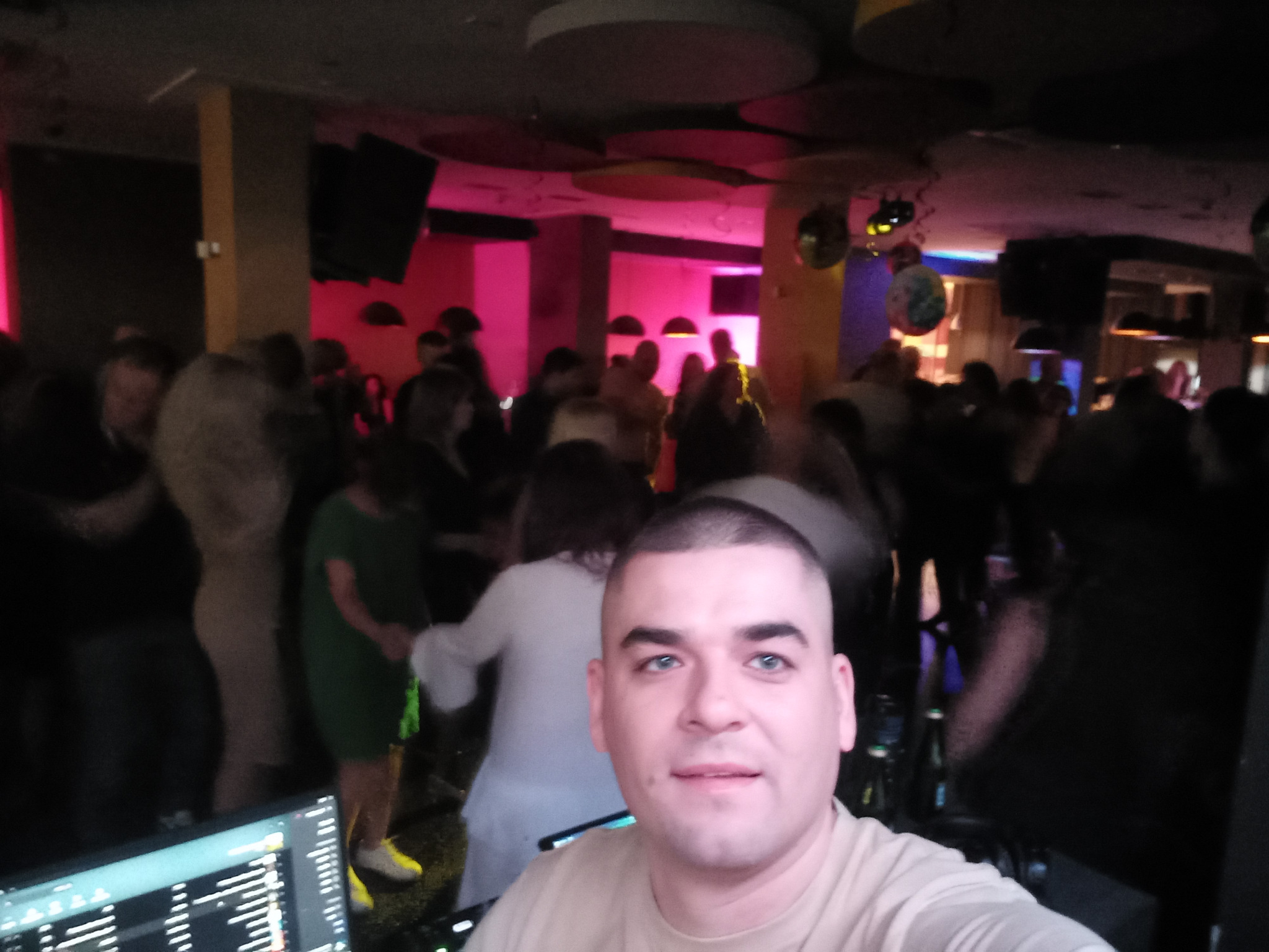 DJ robi selfie na tle roztańczonego tłumu w klubie z dekoracyjnym oświetleniem. Widoczny sprzęt DJ-ski.