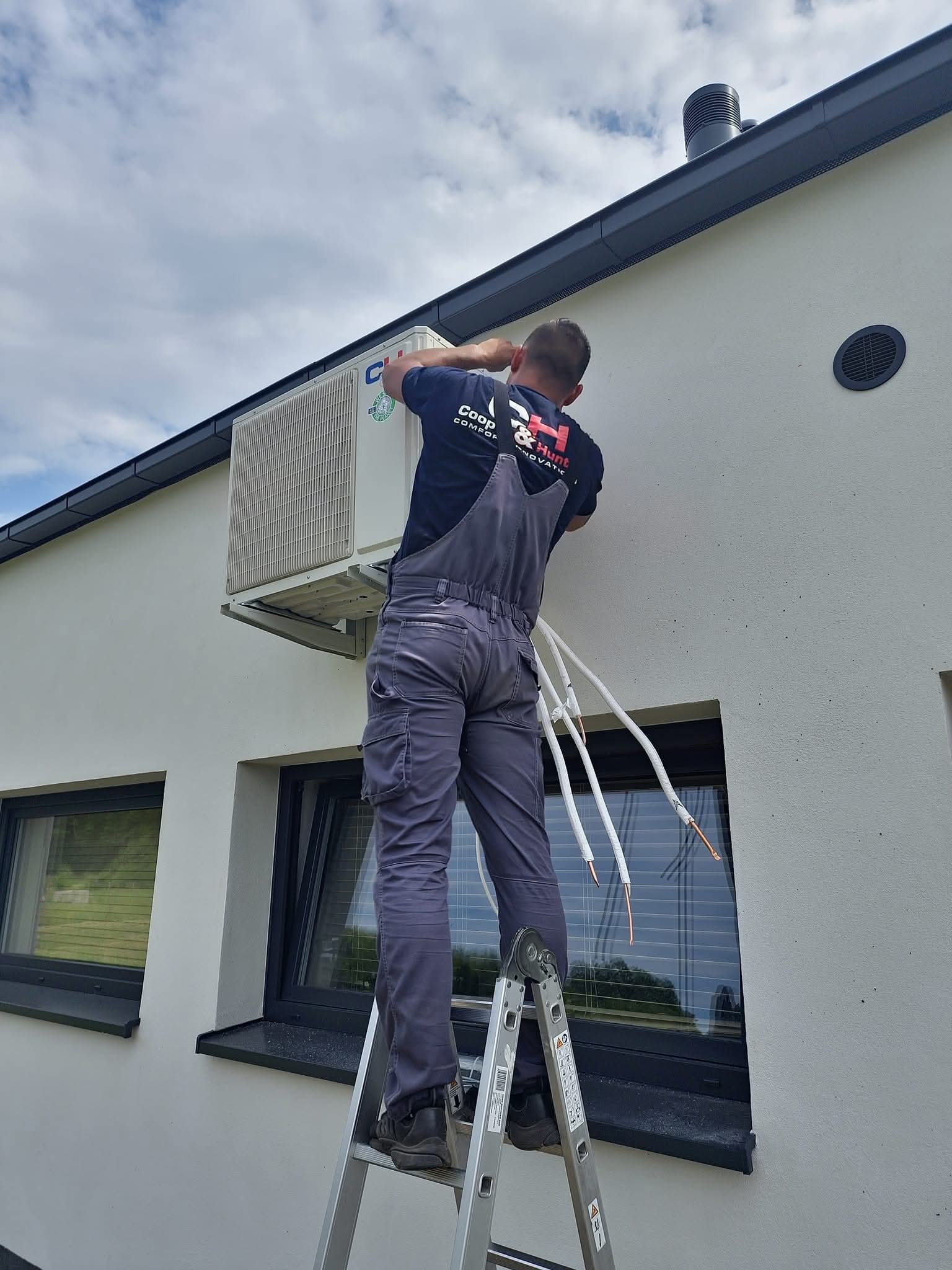 Montaż klimatyzacji zewnętrznej na ścianie domu przez instalatora na drabinie, widoczne rury i jednostka klimatyzatora marki CHIGO.