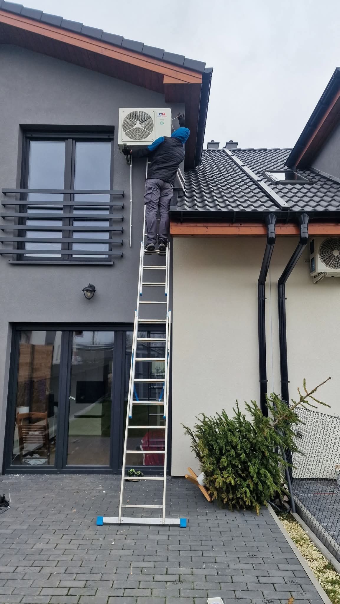 Montaż klimatyzacji na fasadzie domu: mężczyzna na drabinie instaluje jednostkę zewnętrzną, widoczne okna i dach. Szara elewacja, nowoczesny styl.