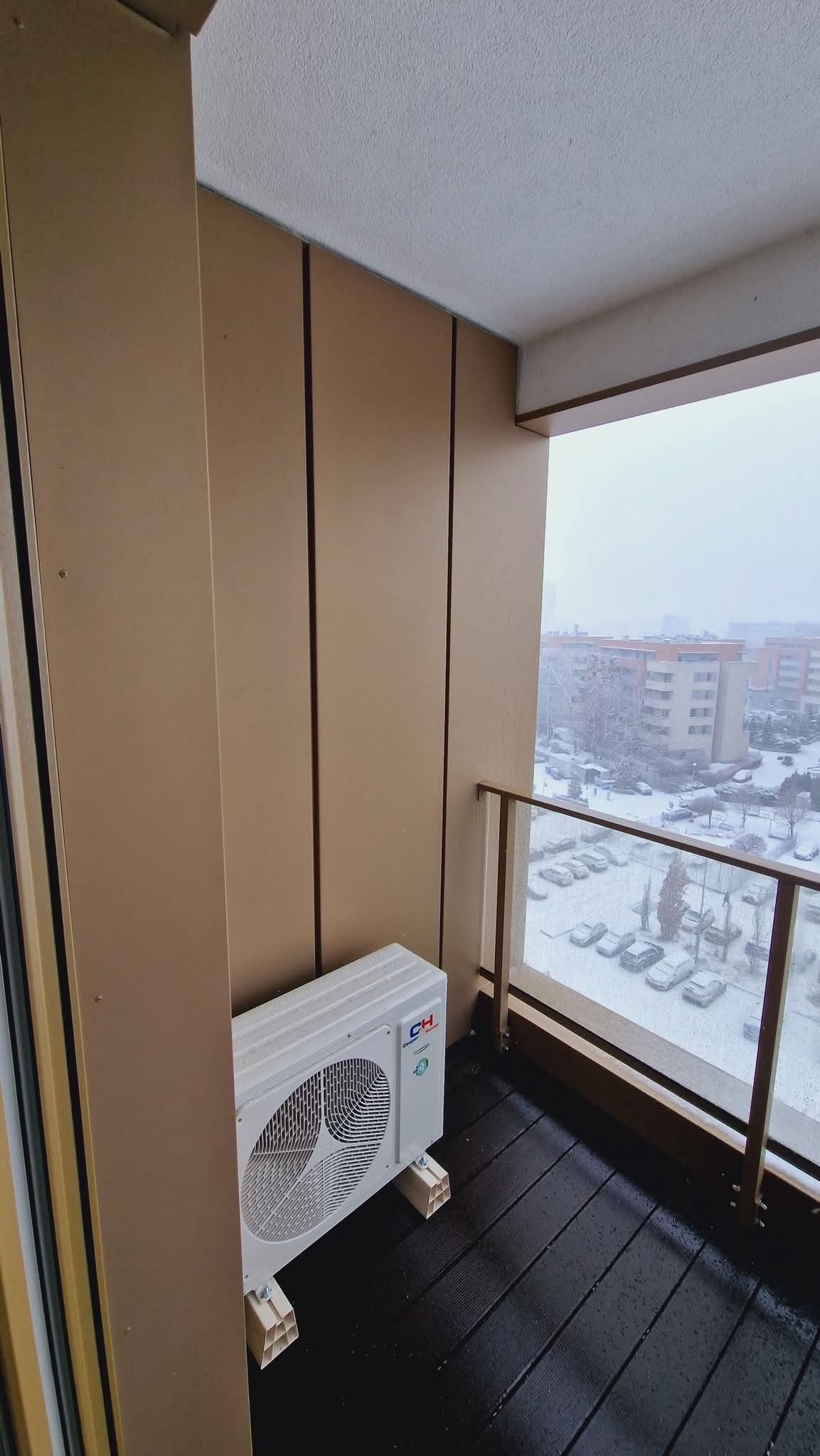 Biała jednostka zewnętrzna klimatyzacji na balkonie z drewnianą podłogą i widokiem na zasypane śniegiem budynki. Montaż klimatyzacji w zimowych warunkach.