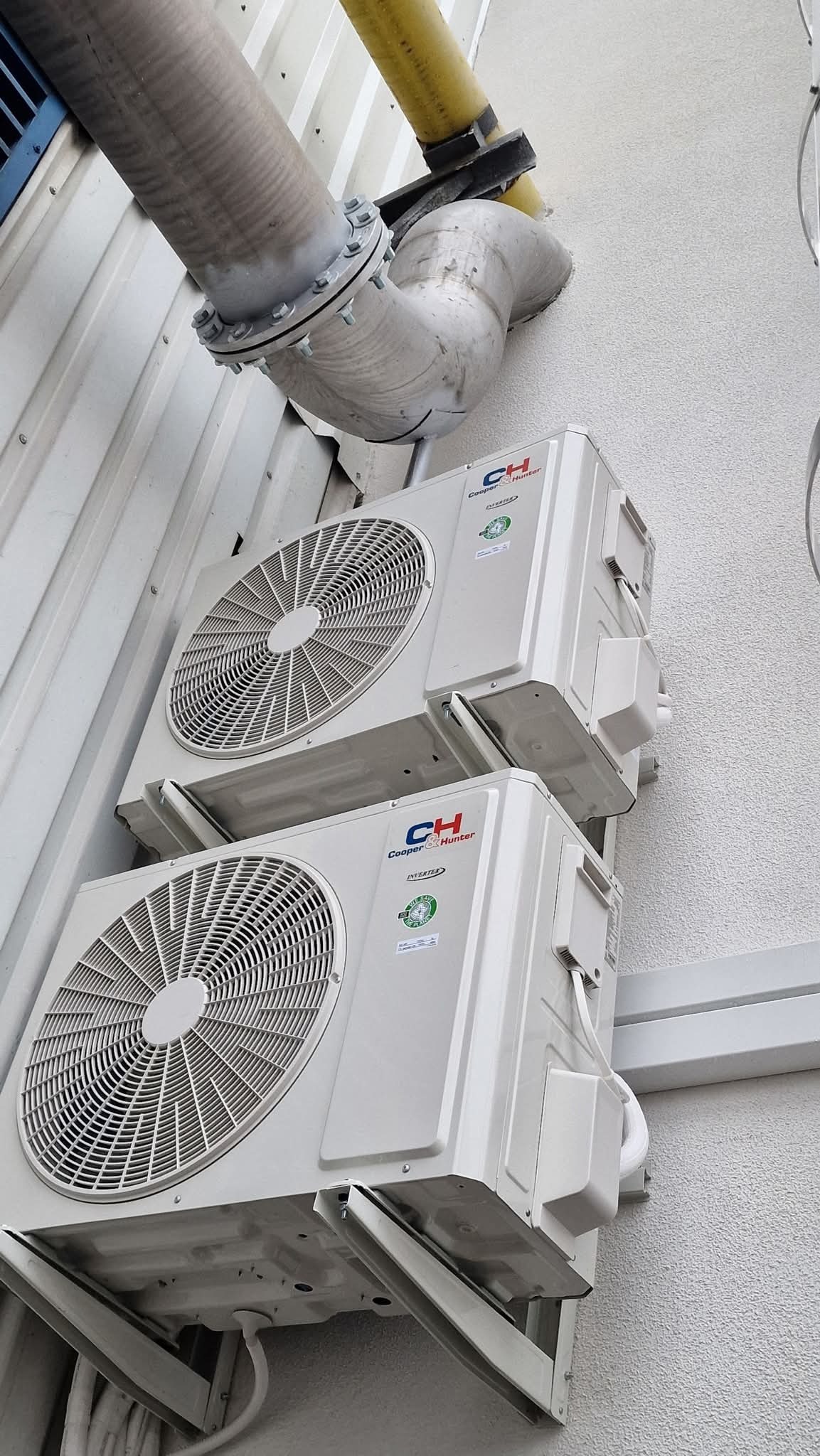 Dwa białe klimatyzatory Cooper&Hunter zamontowane na zewnątrz budynku, z widocznymi rurami instalacyjnymi i elementami mocującymi. Kąt ujęcia z dołu podkreśla instalację.