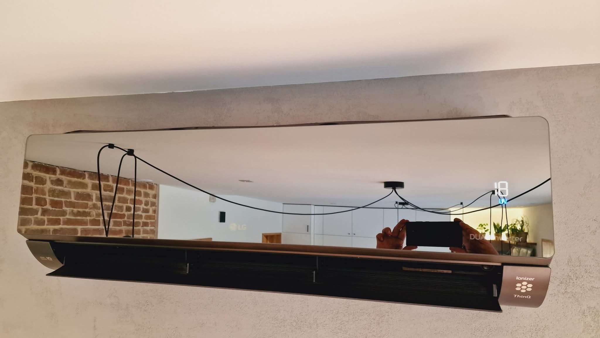 Nowoczesna klimatyzacja LG Dual Inverter zamontowana na ścianie, z widocznym wyświetlaczem temperatury i odbiciem wnętrza pokoju. Widoczne przewody zasilające.