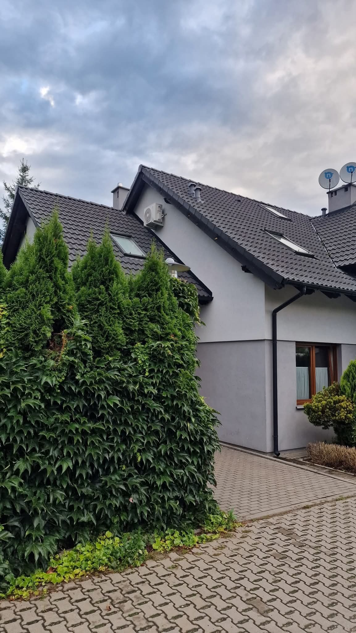 Szary dom z ciemnym dachem, widoczny klimatyzator na ścianie, okna dachowe i ozdobne krzewy w ogrodzie. Na dachu anteny satelitarne.