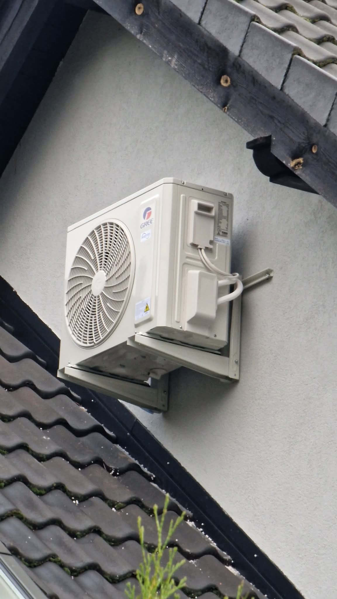 Biała jednostka zewnętrzna klimatyzacji marki Gree, zamontowana na elewacji jasnego budynku z ciemnym dachem, widoczna z boku, z elementami instalacji.