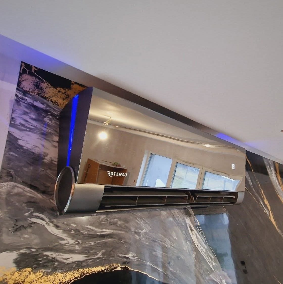 Nowoczesny klimatyzator Rotenso w kolorze srebrnym, zamontowany na ścianie z marmurowym wzorem, z niebieskim podświetleniem LED. Widoczne odbicie wnętrza pokoju.