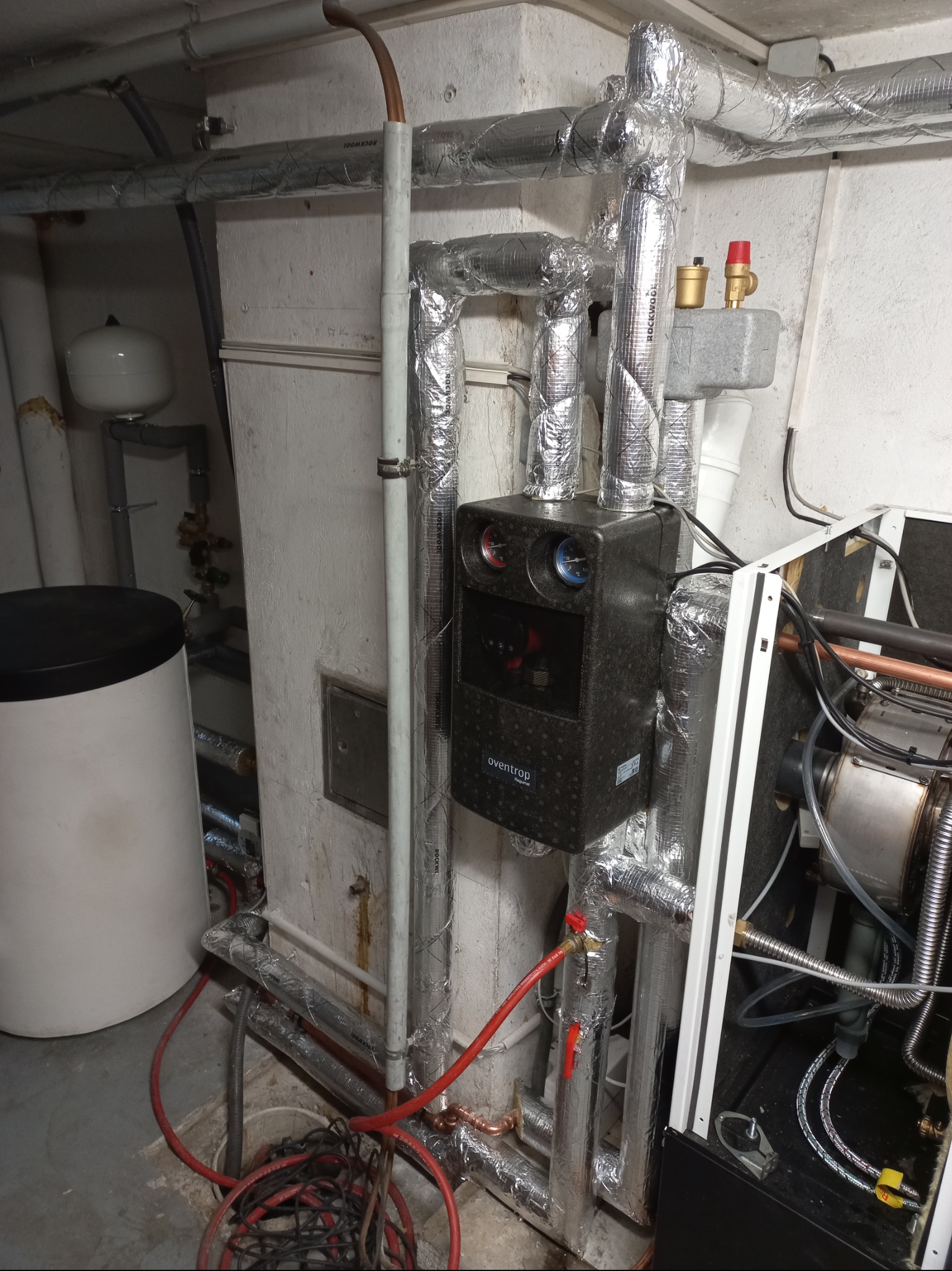 Kotłownia z piecem, zbiornikiem i rurami w izolacji aluminiowej. Widoczne elementy instalacji grzewczej, w tym czarny regulator temperatury Oventrop.