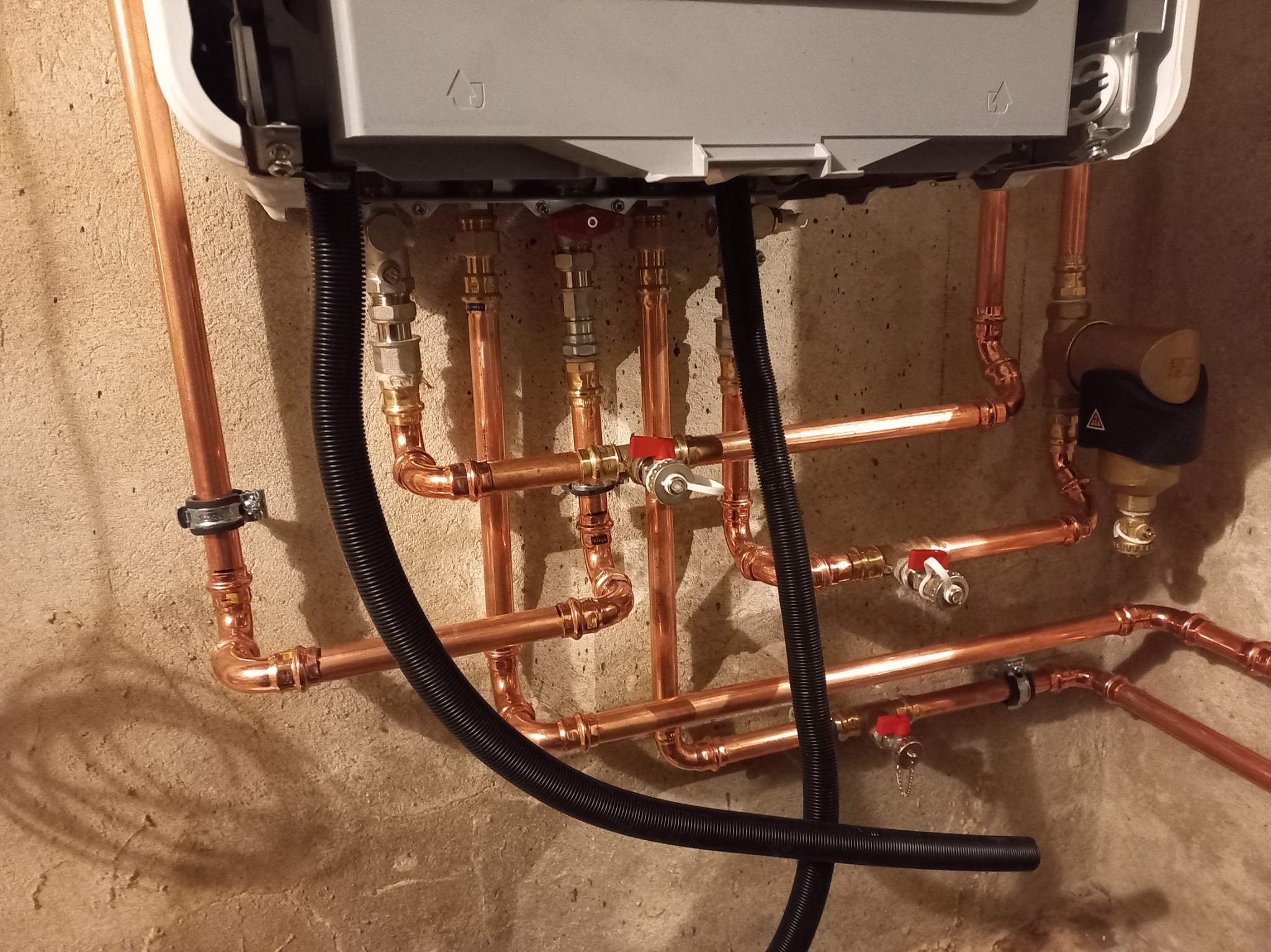 Miedziane rury instalacji grzewczej z zaworami, zamontowane pod piecem gazowym na tle surowej ściany. Widoczne złącza i czarny przewód karbowany.