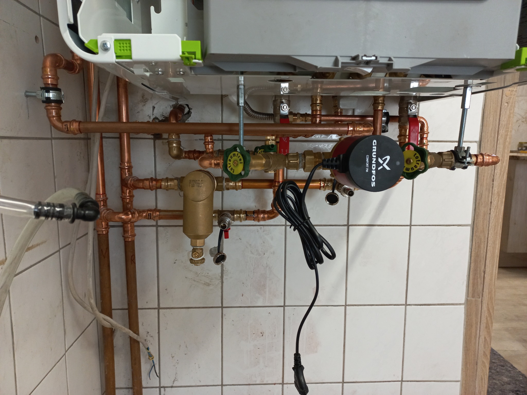 Zamontowany piec gazowy z miedzianą instalacją, widoczna pompa obiegowa Grundfos Comfort PM i zawory. Białe płytki na ścianie.