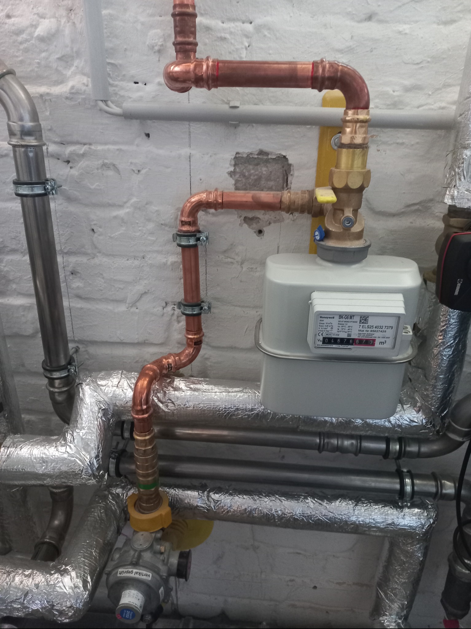Instalacja gazowa z miedzianymi rurami i licznikiem gazu Honeywell BK-G6 MT na tle białej, ceglanej ściany. Widoczne izolowane rury.