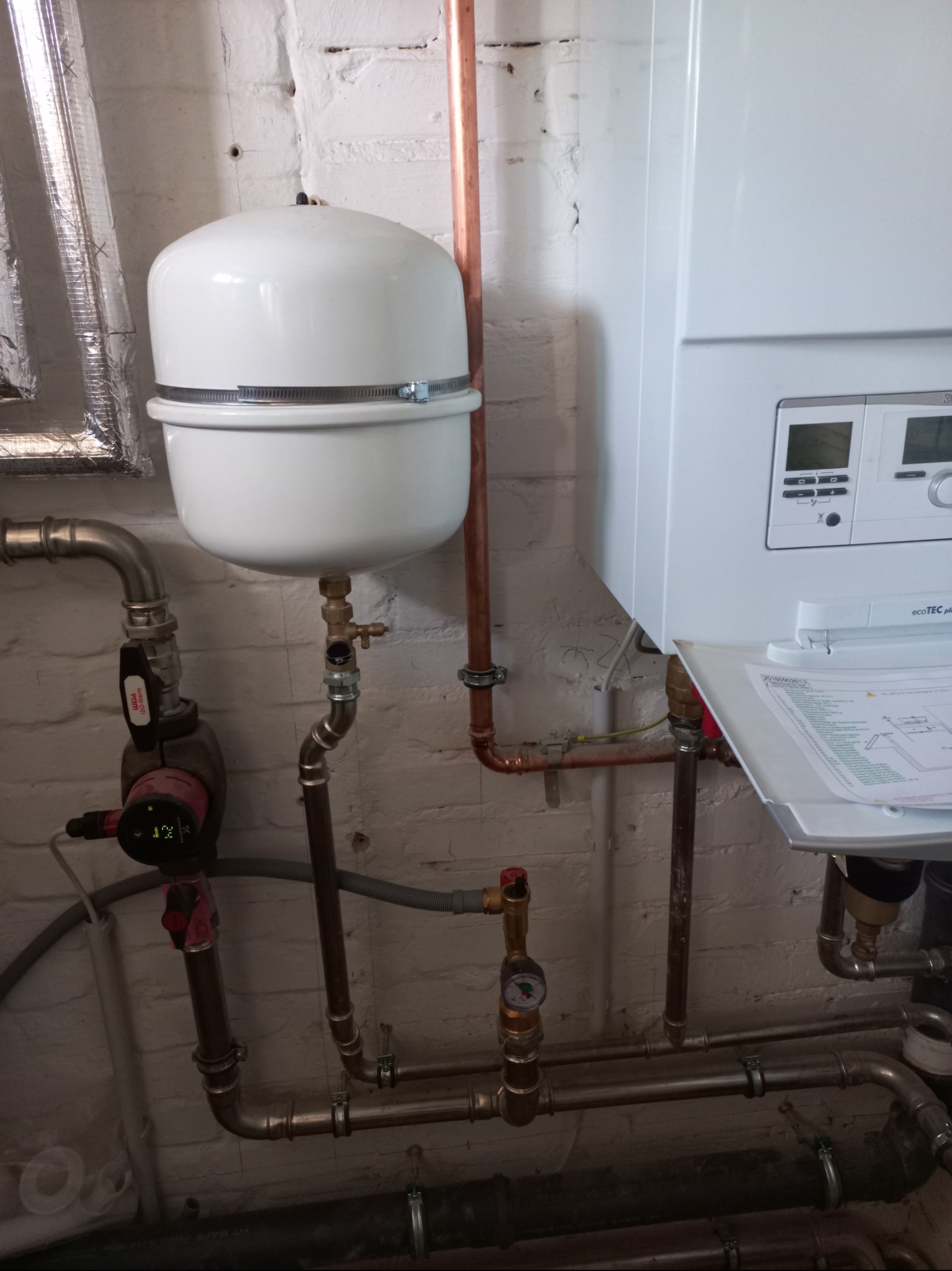 Biały piec gazowy Vaillant Ecotec Plus z podłączonymi miedzianymi rurami, manometrem i pompą obiegową na tle białej, ceglanej ściany piwnicy.