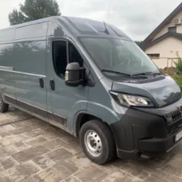 Szary bus dostawczy marki Peugeot, zaparkowany na posesji z kostki brukowej, z domem w tle. Widok z boku, lekko z przodu, na tle zieleni.