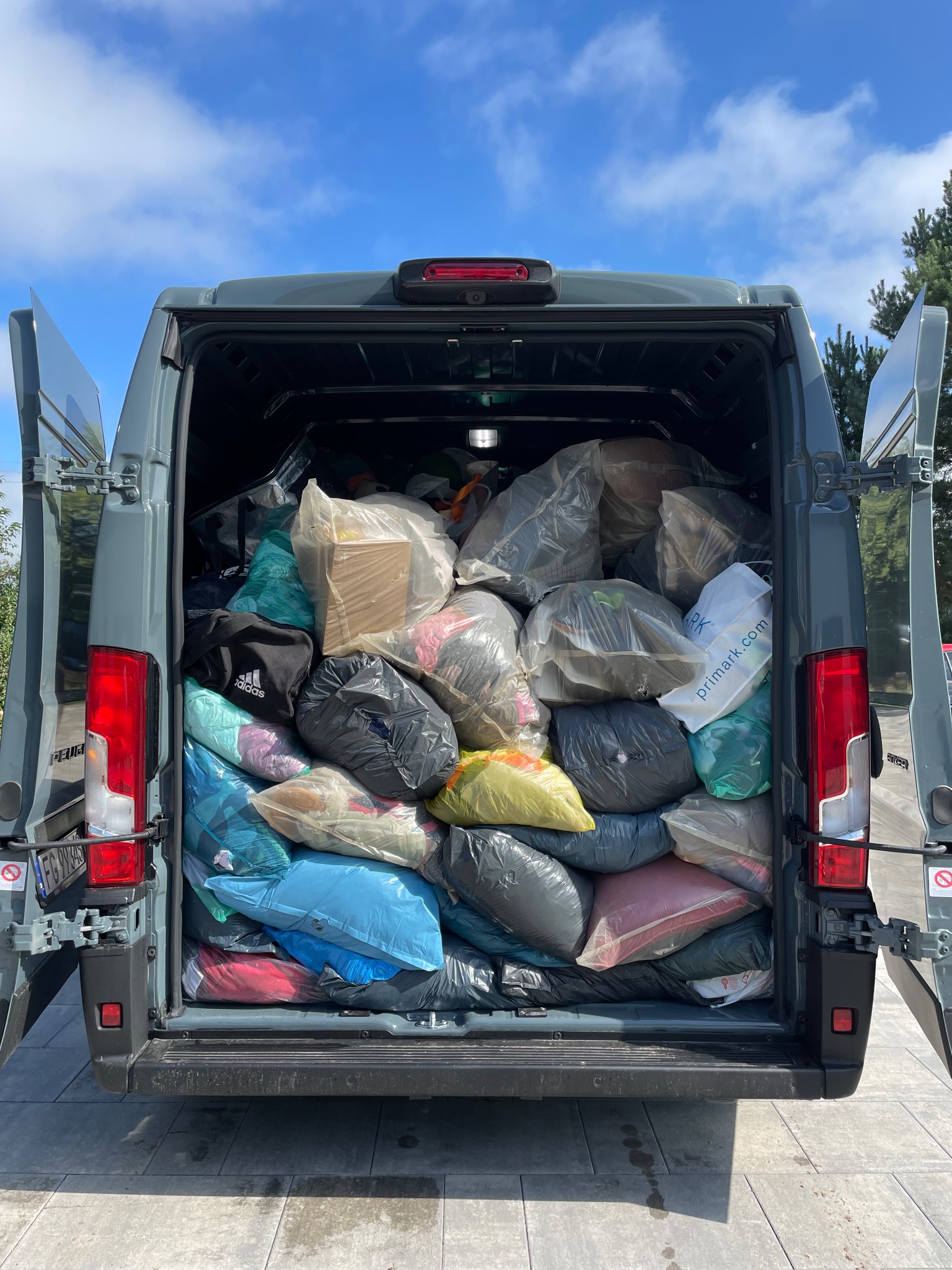Otwarty bagażnik szarego busa wypełniony workami i torbami z odzieżą, widoczne torby marek Adidas i Primark, niebieskie niebo w tle.