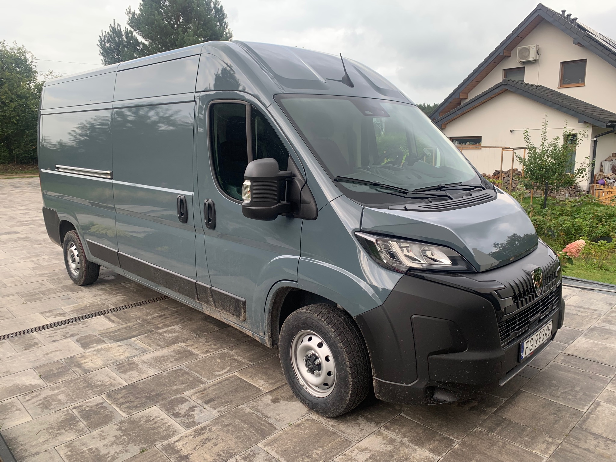 Szary bus dostawczy marki Peugeot, zaparkowany na posesji z kostki brukowej, z domem w tle. Widok z boku, lekko z przodu, na tle zieleni.