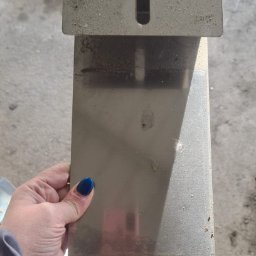 KD PRODUCTS Klaudia Gniazdowska - Szlifowany element metalowy z otworami, trzymany w dłoni. Widoczny fragment wózka warsztatowego w tle. Detal konstrukcyjny z blachy nierdzewnej.