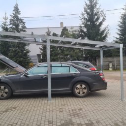 KD PRODUCTS Klaudia Gniazdowska - Metalowa konstrukcja zadaszenia dla samochodów, szara, na posesji z kostki brukowej. W tle budynek i drzewa iglaste.