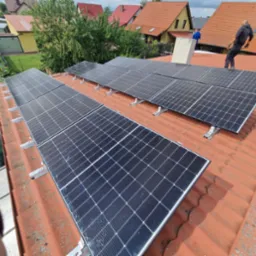 Panele słoneczne na dachu z czerwonej dachówki, widok z góry. Instalacja w trakcie montażu, pracownik na dachu. W tle zabudowa jednorodzinna i zieleń.