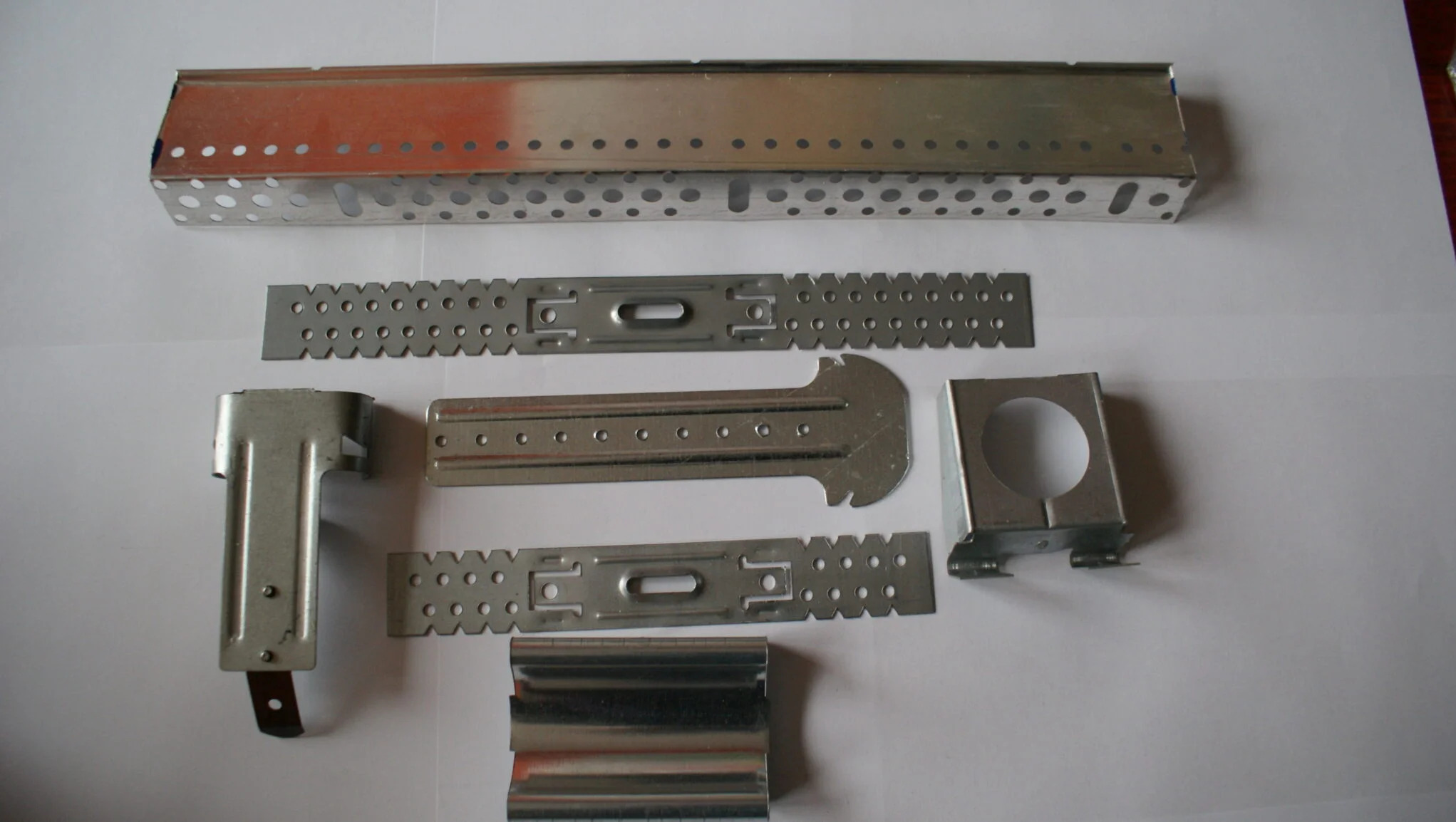 Zestaw metalowych elementów z otworami, prawdopodobnie do montażu lub konstrukcji, ułożonych na białym tle. Różne kształty i rozmiary.