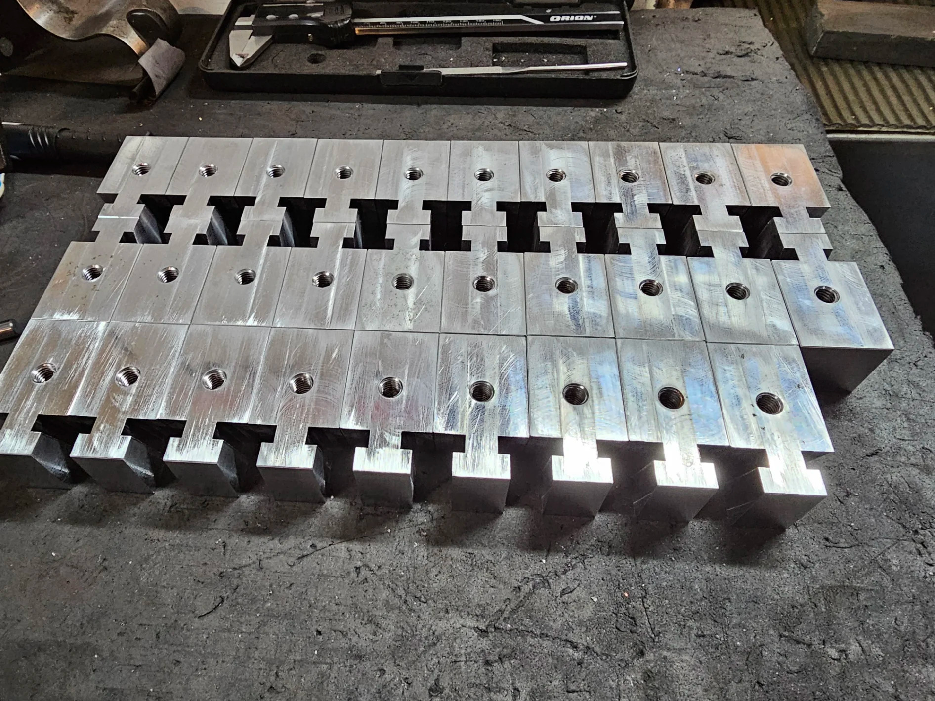 Precyzyjnie wycięte, metalowe elementy z otworami, ułożone rzędami na szorstkiej powierzchni. Widoczne narzędzia pomiarowe w tle sugerują etap kontroli jakości.