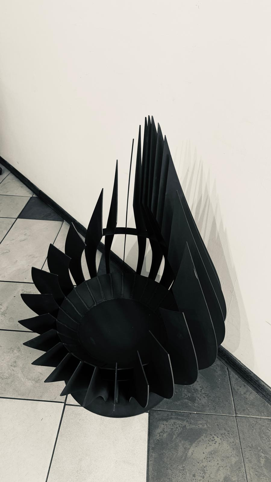 Czarna, metalowa konstrukcja o spiralnym kształcie z ostrymi krawędziami, ustawiona na podłodze z płytek w odcieniach szarości i bieli, przy białej ścianie.